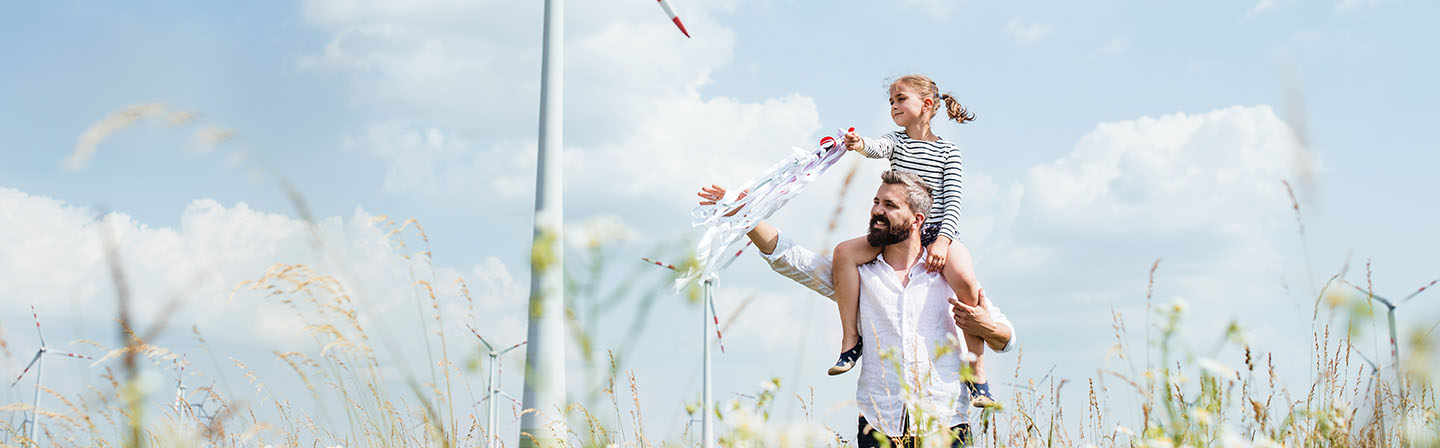 A father walks with his daughter across a field in a wind farm.