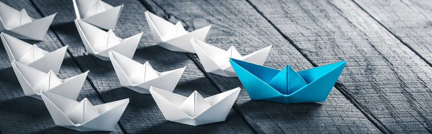 Blue Paper Boat Leading A Fleet Of Small White Boats On Wooden Table